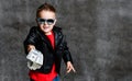 Smiling rich kid boy millionaire in sunglasses, leather jacket and red t-shirt handing us a bundle stack of dollars cash