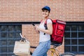 Smiling rider woman wearing red backpack delivering food on a bike, checking order with smart phone while standing on street in Royalty Free Stock Photo