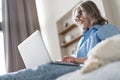 Smiling 50s mature woman sitting on sofa, using laptop, working, chatting, spending time in social media