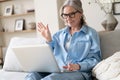 Smiling 50s mature woman sitting on sofa, using laptop, working, chatting, spending time in social media