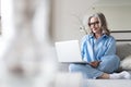 Smiling 50s mature woman sitting on sofa, using laptop, working, chatting, spending time in social media