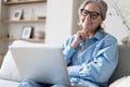 Smiling 50s mature woman sitting on sofa, using laptop, working, chatting, spending time in social media