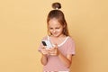Smiling satisfied dark haired little girl wearing striped T-shirt isolated over beige background using smartphone playing video Royalty Free Stock Photo