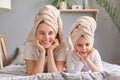 Smiling satisfied mother and daughter laying in bed wearing towel posing in bedroom at home looking at camera holding chins