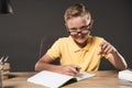 smiling schoolboy in eyeglass pointing by finger and doing homework at table with books colour pencils and textbook on grey