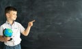 a smiling schoolboy at school with a globe stands in front of a chalk board and points his finger at a place for text Royalty Free Stock Photo