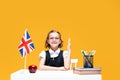 Smiling schoolgirl raising hand sitting at the desk during lesson. English lesson. Great Britain flag