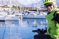 Smiling senior adult man with yellow helmet looks at the camera while passing with the bicycle near the port. Sailboats in the Royalty Free Stock Photo