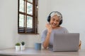 Smiling senior asian woman with headphones on her head sitting at a table in front of a laptop and greeting family Royalty Free Stock Photo
