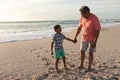 Smiling senior biracial man holding hand while looking at grandson standing on shore against sky