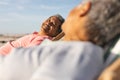 Smiling senior biracial man sitting on chair while looking at woman relaxing on beach Royalty Free Stock Photo