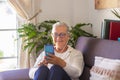 Smiling senior lady sitting on sofa at home using her smart phone for chatting with friends or family - woman white hair, glasses Royalty Free Stock Photo