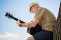 Smiling senior man in hat leaning at tree and playing guitar Royalty Free Stock Photo