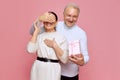 Smiling senior man, husband covering woman's eyes, greeting with holiday and presenting gift against pink studio