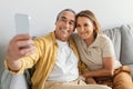 Smiling senior man and lady taking selfie on smartphone, resting on sofa at home and smiling at webcamera