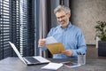 Smiling senior man sitting at home at work desk with laptop, opening and reading received letter Royalty Free Stock Photo