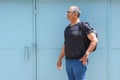 Smiling senior man standing in front of blue door in a city holding his black backpack, looking away to side. Royalty Free Stock Photo