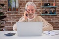 Smiling senior man using laptop computer and cellphone at home. Brick`s wall on background