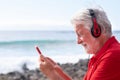 Smiling senior man with white hair and beard having video conversation on mobile phone. Sitting on the rock beach facing the sea Royalty Free Stock Photo