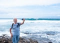 Smiling senior man with white hair and beard takes a selfie with the large waves behind him. Horizon over water in background. Royalty Free Stock Photo