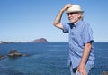 Smiling senior man white haired wearing a straw hat standing and enjoying vacation and freedom in seascape. Blue sea red mountain Royalty Free Stock Photo