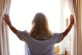 Smiling senior white woman opening the curtains on a sunny morning, back view, waist up