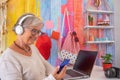 Smiling senior woman with headphones using laptop computer talking on mobile phone. Camera and sea decoration on background Royalty Free Stock Photo
