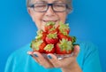 Smiling senior woman holding plate of fresh ripe strawberries. Blue background