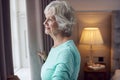 Smiling Senior Woman At Home Wearing Pyjamas Opening Bedroom Curtains And Looking Out Of Window