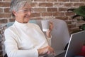 Smiling senior woman sitting at home using laptop computer holding a tea cup - brick wall background Royalty Free Stock Photo