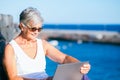 Smiling senior woman sitting outdoor at sea port using laptop computer. Relaxed elderly woman wearing sunglasses, sunset light