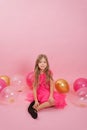 Smiling seven-year-old girl dressed in a fashionable dress sits on a pink background surrounded by balloons. Celebrate your