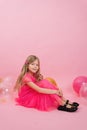 Smiling seven-year-old girl dressed in a fashionable dress sits on a pink background surrounded by balloons. Celebrate your