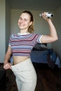 Smiling slim woman in big pants with dumbbell in hand in apartment. Royalty Free Stock Photo