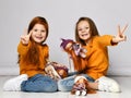 Smiling small girls sisters in yellow longsleeves, jeans and sneakers sitting on floor playing with dolls and showing