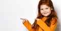 Smiling small red haired girl with freckles in yellow comfortable longsleeve standing and pointing aside with fingers