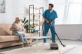 smiling social worker cleaning carpet with vacuum cleaner and looking at senior woman