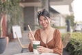 A smiling Southeast Asian woman sitting outside the coffee house places the straw inside her drink