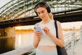 Smiling sportive woman in headphones using mobile phone while standing on embankment