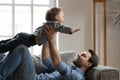 Smiling strong dad lying on comfortable sofa, lifting small son.