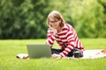 Smiling student girl in earphones using laptop while sitting on blanket on green grass in city park Royalty Free Stock Photo