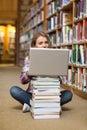 Smiling student sitting on library floor using laptop on pile of books