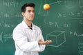 Smiling student with white coat throwing an apple up, on green chalkboard with equations background.