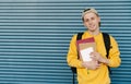 Smiling student in a yellow sweatshirt and cap stands on the background of a blue wall with books and notebooks and a cup of Royalty Free Stock Photo