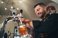 Smiling stylish bearded barman dressed black uniform with an apron tapping fresh lager beer into glass mug at bar counter. Royalty Free Stock Photo