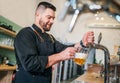 Smiling stylish bearded barman dressed black uniform with an apron tapping fresh lager beer into glass mug at bar counter. Royalty Free Stock Photo