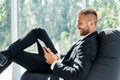 Smiling successful businessman in elegant suite using tablet sitting on bean bag chair in creative office Royalty Free Stock Photo