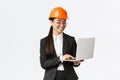 Smiling successful female asian industrial engineer, factory manager in safety helmet and business suit using laptop Royalty Free Stock Photo