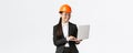 Smiling successful female asian industrial engineer, factory manager in safety helmet and business suit using laptop Royalty Free Stock Photo