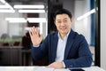 Smiling and successful young Asian businessman in business suit sitting at office desk, looking at camera, greeting and Royalty Free Stock Photo